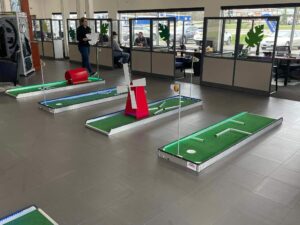 Portable mini golf setup in a car dealership showroom, featuring multiple green courses, a red obstacle, and players engaging in family-friendly fun.