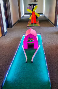 Portable LED mini golf course setup featuring brightly colored obstacles, including a pink structure and a windmill, designed for corporate team-building events in an office environment.