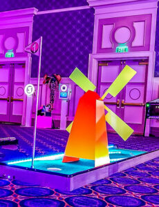 Colorful LED mini golf hole featuring a windmill obstacle, illuminated in vibrant hues, set in a stylish event space, promoting fun portable mini golf experiences for events.