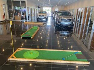 Portable mini golf course setup in a car dealership showroom with two putting greens, golf clubs, and vehicles in the background, enhancing family-friendly atmosphere and customer engagement.
