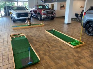 Portable mini golf course setup in a car dealership showroom featuring synthetic grass, golf clubs, and holes, enhancing family-friendly atmosphere and customer engagement.